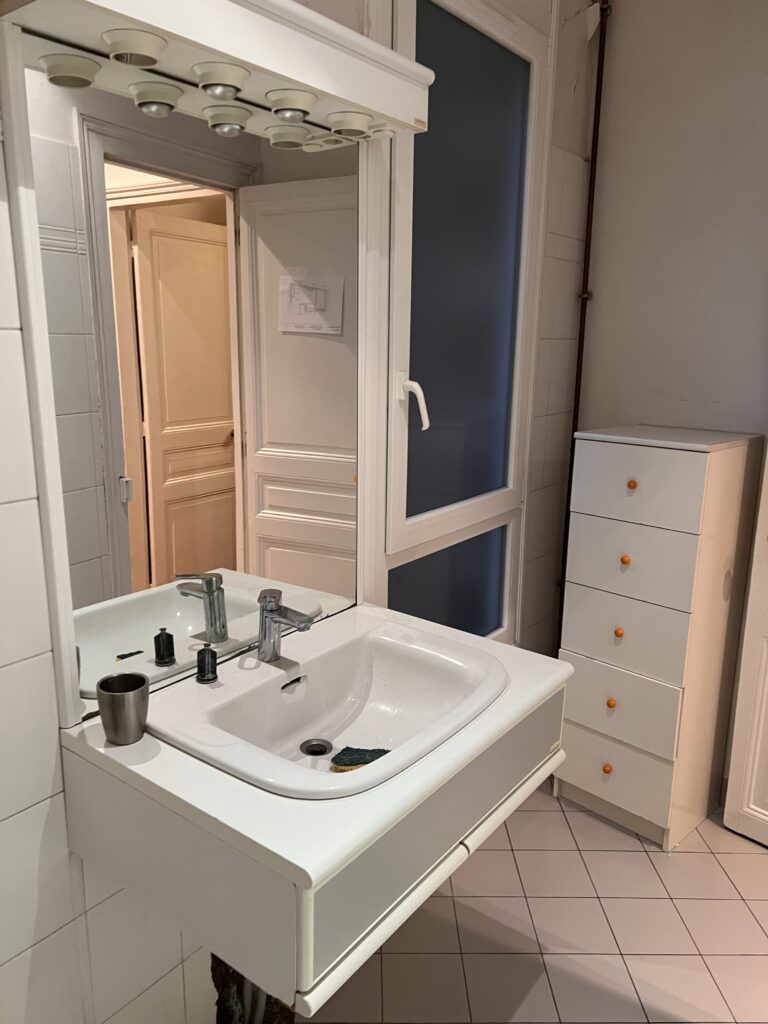 Vue de la salle de douche avant rénovation, avec un lavabo suspendu, un grand miroir et une fenêtre apportant de la lumière naturelle.