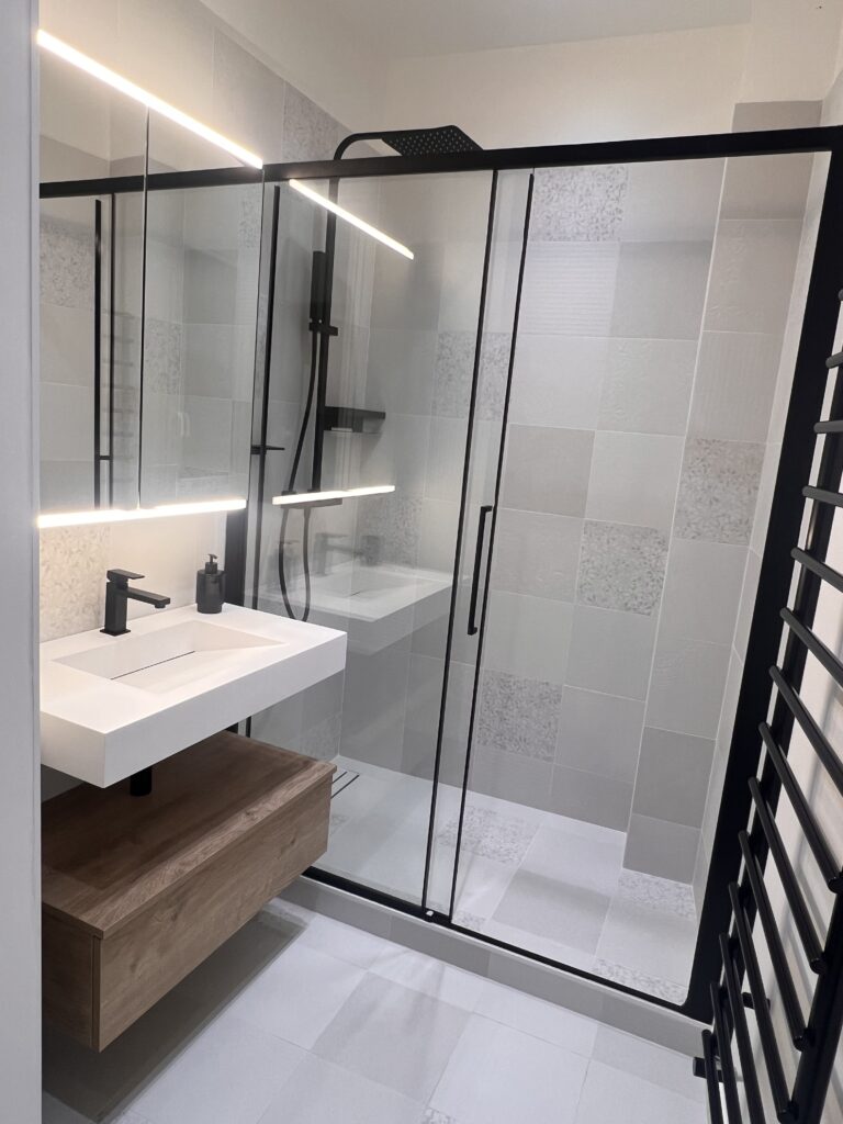Vue complète d’une salle de douche rénovée à Saint-Mandé avec meuble vasque suspendu, paroi vitrée noire et carrelage texturé beige