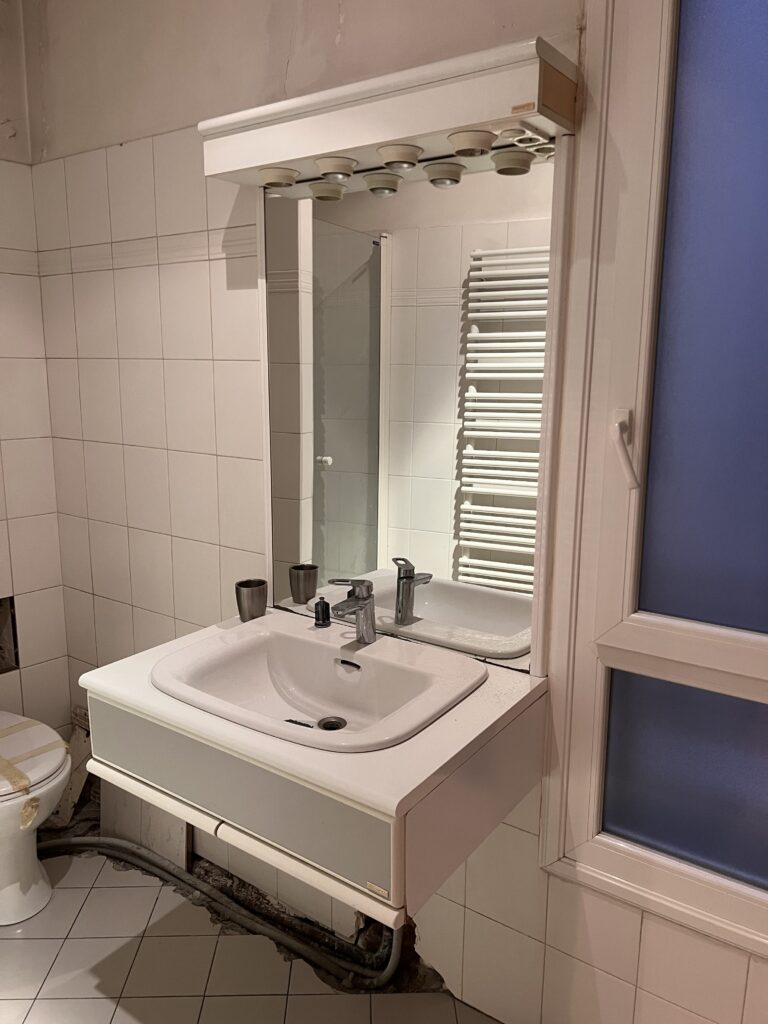 Vue d’un meuble vasque suspendu avec miroir et spots lumineux avant rénovation, dans une salle de douche à Saint-Mandé.