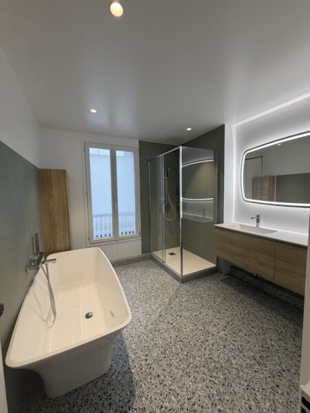 Vue d’ensemble d’une salle de bains rénovée avec baignoire îlot, douche vitrée, meuble vasque suspendu et sol en terrazzo